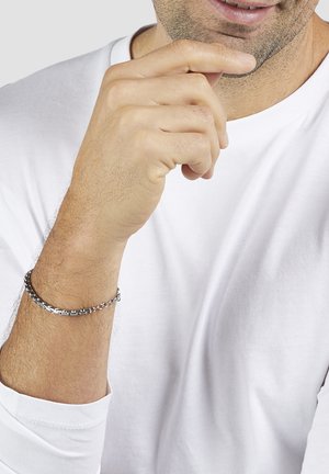 Man in white long-sleeve shirt touching chin with right hand, wearing a silver chain bracelet on left wrist, smiling slightly.