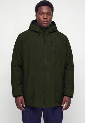Man wearing a dark green hooded jacket with front zipper and zippered chest pocket, standing against a plain light background.
