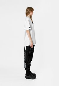 Camiseta blanca de gran tamaño con detalles en negro, combinada con pantalones cargo negros que presentan patrones gráficos y cremalleras. Unos zapatos moldeados negros completan el look.