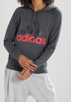 Person iført en mørkegrå hættetrøje med et sort og rødt Adidas-logo, kombineret med lysegrå teksturerede bukser, stående med armene krydset.