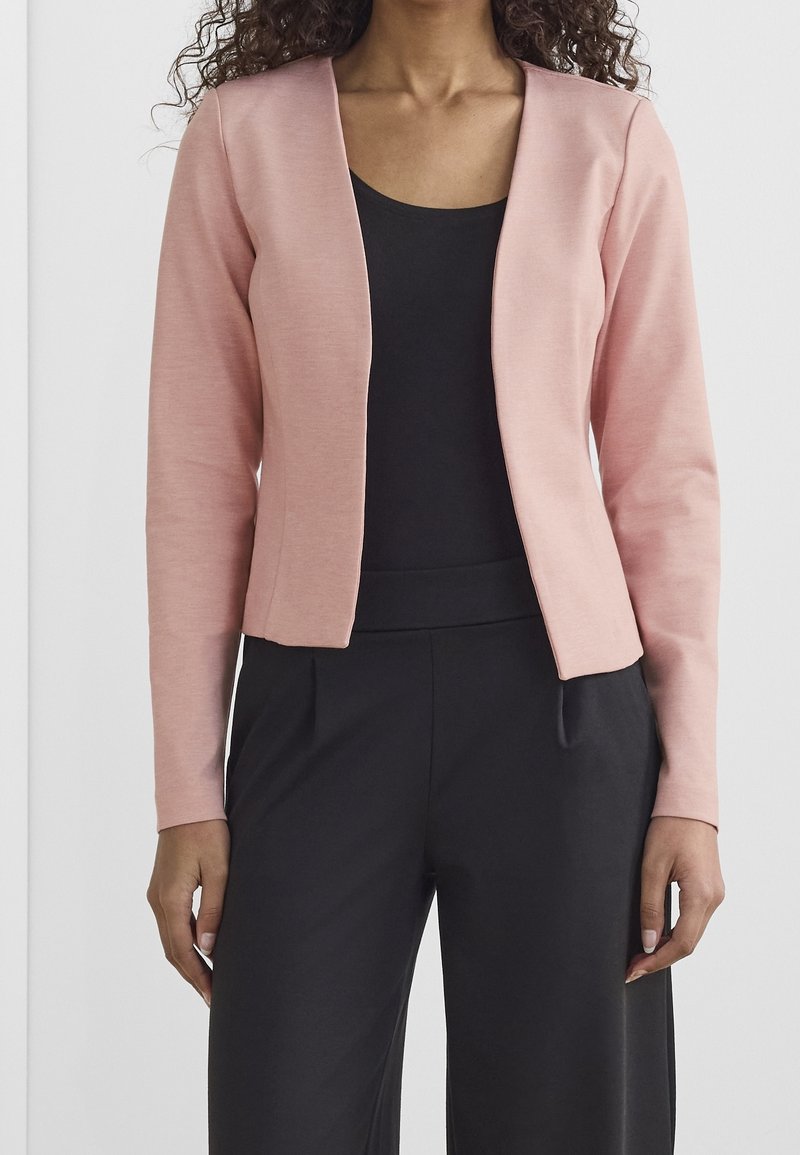Blazer ajusté rose clair avec une texture lisse et un design à devant ouvert. Associé à un haut noir et un pantalon noir taille haute.