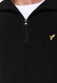 Juodas vilnos džemperis su užtrauktuku priekinėje dalyje, kairėje pusėje su mažu auksiniu erelio siuvinėjimu ir aukšta apykakle.