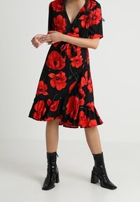 Černé květinové zavinovací šaty s červenými mákovými vzory, krátkými rukávy a volánem na lemu, spárované s černými kotníkovými botami.