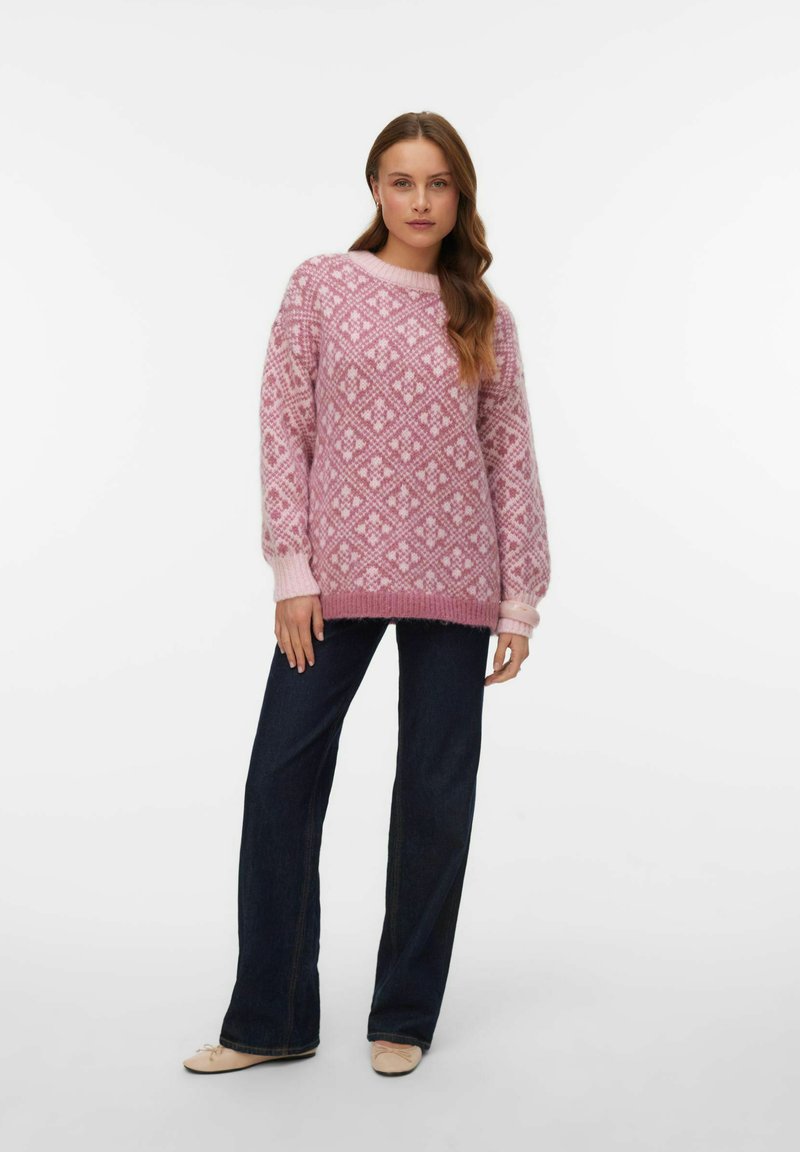 Pull en tricot rose clair avec un motif en diamant. Il a un col rond et une coupe décontractée. Associé à un jean flare noir et des ballerines.