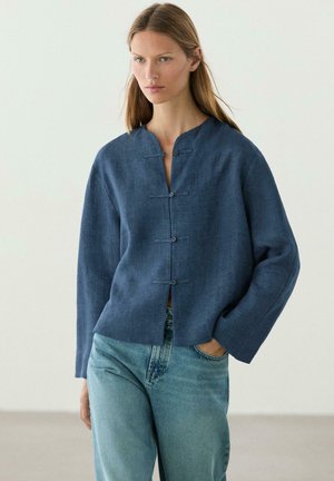 Frau trägt eine lockere dunkelblaue Bluse mit Knöpfen und langen Ärmeln sowie hellblaue Jeans, steht mit einer Hand in der Tasche vor einem einfarbigen Hintergrund.