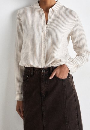 Vrouw met een beige-wit gestreept shirt met verticale strepen en lange mouwen, ingestopt in een high-waisted donkerbruine rok, met één hand in de zak van de rok.