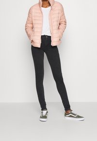 Chaquetón acolchado rosa claro, jeans negros ajustados y zapatillas verdes. La chaqueta tiene paneles acolchados horizontales y un cuello alto.