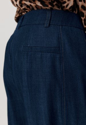 Ciemnoniebieskie dżinsy o dopasowanym kroju, czystych liniach, subtelnym haftowaniu i tylniej kieszeni. Materiał ma gładką teksturę.