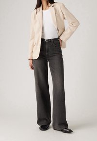 Blazer de textura beige sobre una camisa blanca, combinado con unos vaqueros negros de cintura alta. Los detalles incluyen un cinturón con una hebilla brillante y un corte de pierna ancha.
