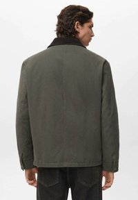 Uomo con capelli scuri e mossi di spalle, che indossa una giacca verde oliva con colletto scuro e jeans neri, su uno sfondo semplice e chiaro.