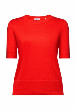 Personne portant un pull rouge à manches courtes, un pantalon noir, un collier chaîne en or, portant un sac à épaule noir, et de longs cheveux tressés.