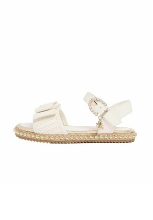 Sandale blanche plate avec bride avant tissée, boucle ornée de perles, bordure de perles dorées le long de la semelle, et petit pendentif en forme de pompon sur la bride de cheville.