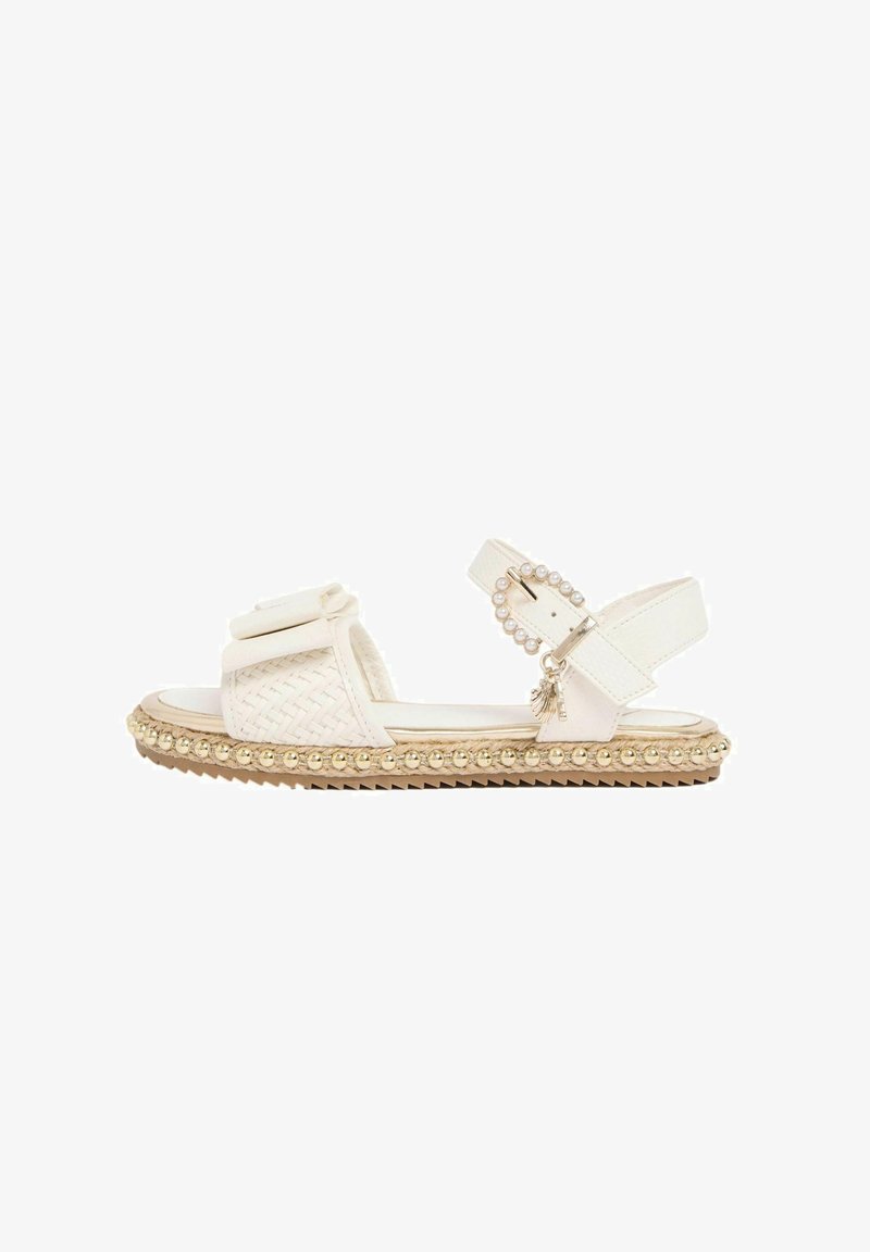 White flat sandal with woven front strap, pearl-adorned buckle, gold bead trim along sole, and small tassel charm on ankle strap.