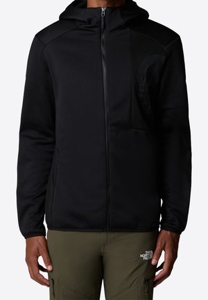 Mężczyzna noszący czarną kurtkę z kapturem na zamek błyskawiczny i oliwkowe spodnie z logo The North Face na lewej udzie, stojący na jednolitym tle.