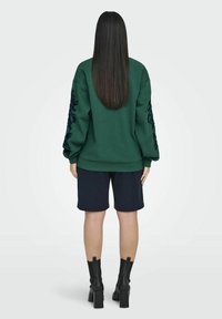 Sudadera verde con mangas extragrandes y bordados florales, combinada con pantalones cortos azul marino y botines negros de tacón, vista desde atrás.