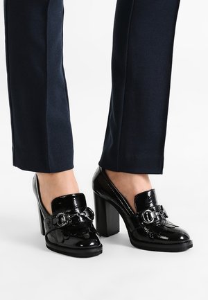 Mocasines de charol negro de tacón alto con detalle de hebilla, usados con pantalones azul marino oscuro sobre un fondo blanco.