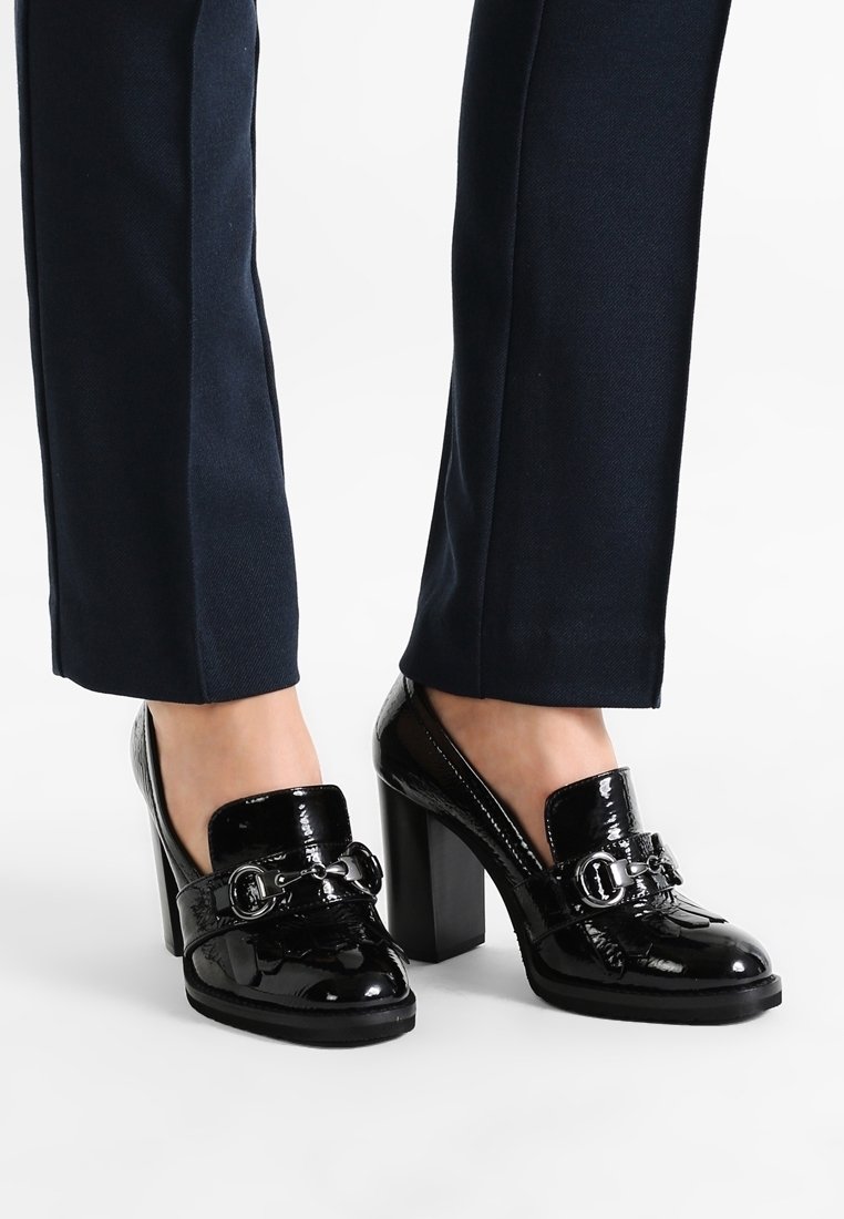 Mocassins à talons hauts en cuir verni noir avec détail boucle portés avec un pantalon bleu marine foncé sur fond blanc.