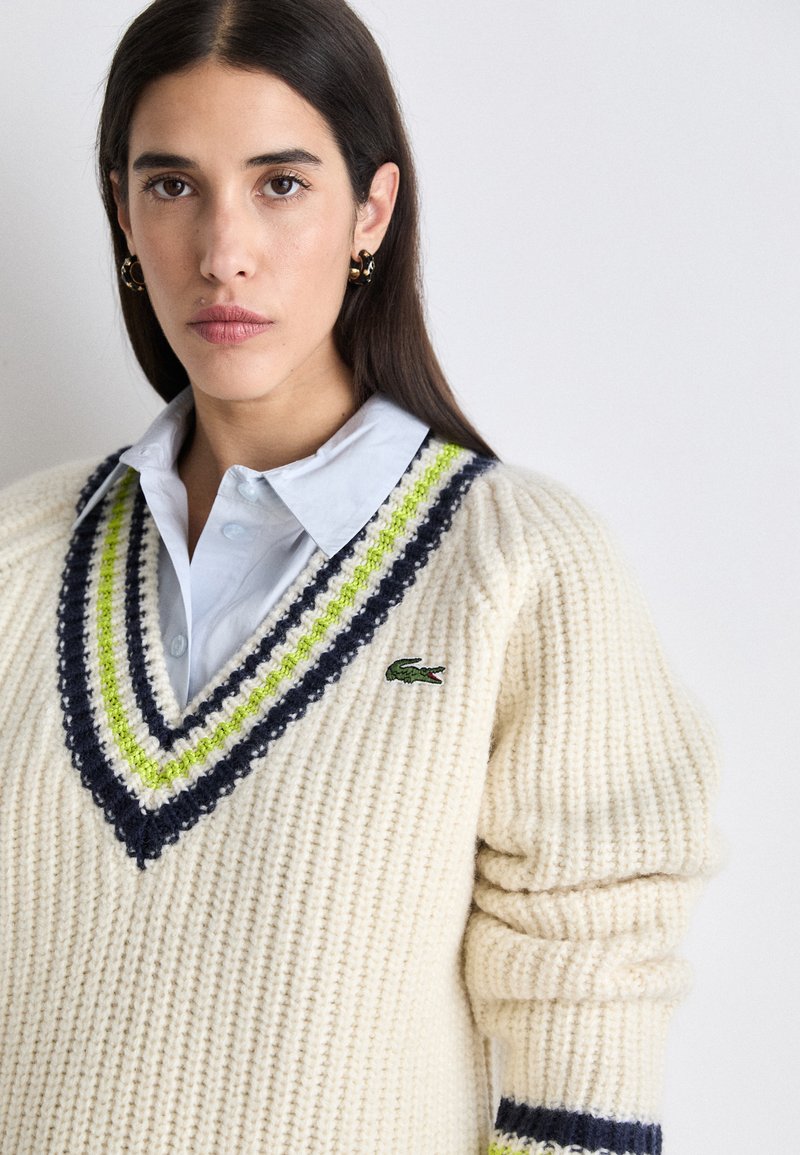 Pull en tricot crème avec décolleté en V, présentant des accents rayés bleu marine et vert lime ainsi qu'un petit logo brodé. Design texturé et épais.