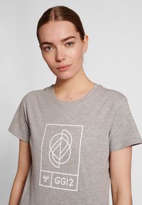 Grå bomullströja med en vit geometrisk logotyp med sammanflätade former och texten "GGI2" nedanför, rund krage, korta ärmar.