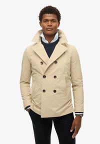 Beige dubbele rijgjas met grote bruine knopen, geknipte revers en zijzakken. Draag het over een marineblauwe trui en een lichtblauw overhemd.