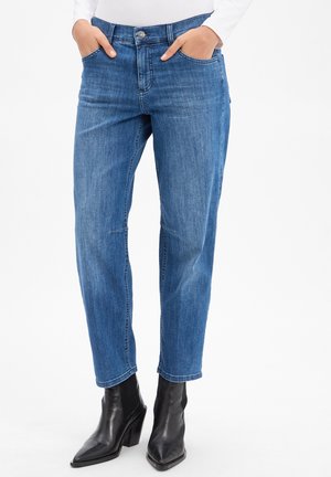 Osoba w niebieskich dżinsach o średnim stanie i prostych nogawkach oraz czarnych botkach z zaokrąglonym noskiem, ręce w kieszeniach na białym tle.