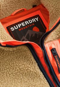 Fleece bunda s hnědou texturovanou podšívkou, oranžovými detaily a černou nášivkou s horským motivem a textem "SUPERDRY MOUNTAIN".
