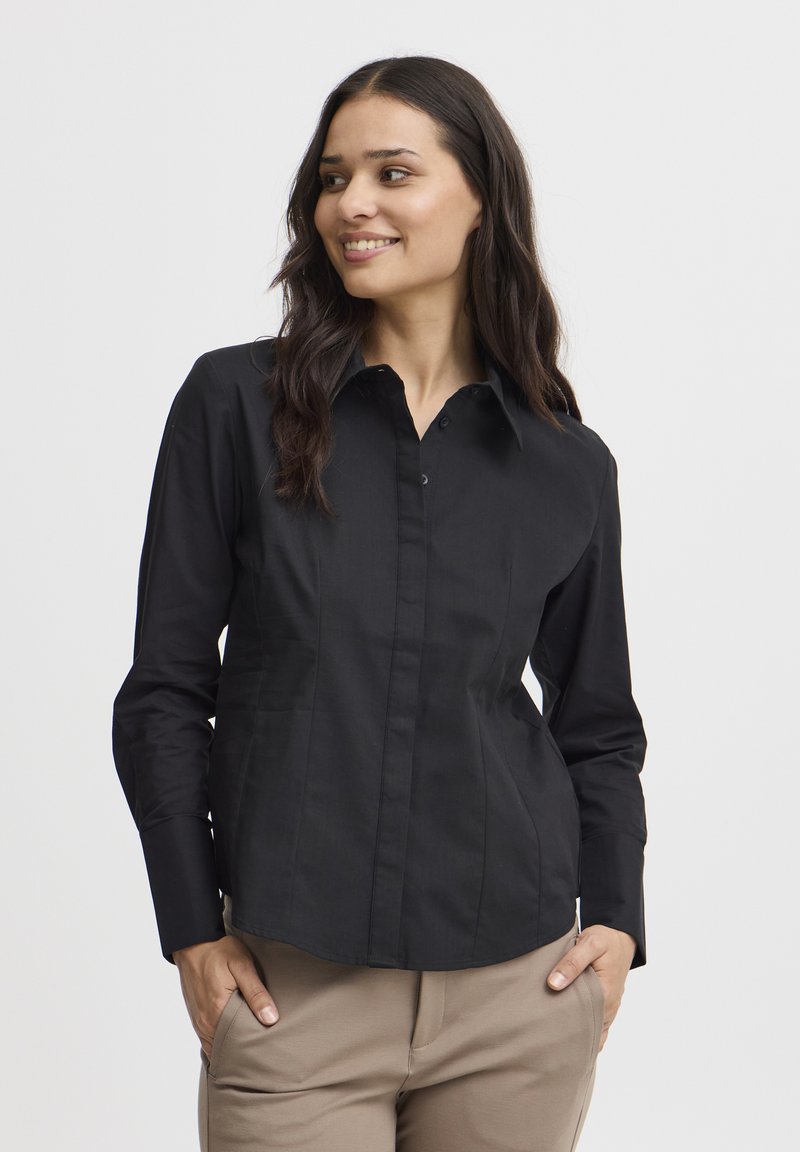 Camicia nera a maniche lunghe con bottoni, dal design aderente, colletto a punta e cuciture visibili. Indossata con pantaloni beige.