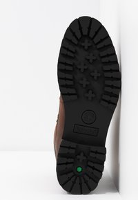 Semelle de botte en cuir marron avec de profonds motifs de semelle noirs. Présente un logo circulaire au niveau du talon et un élément d'accent vert.