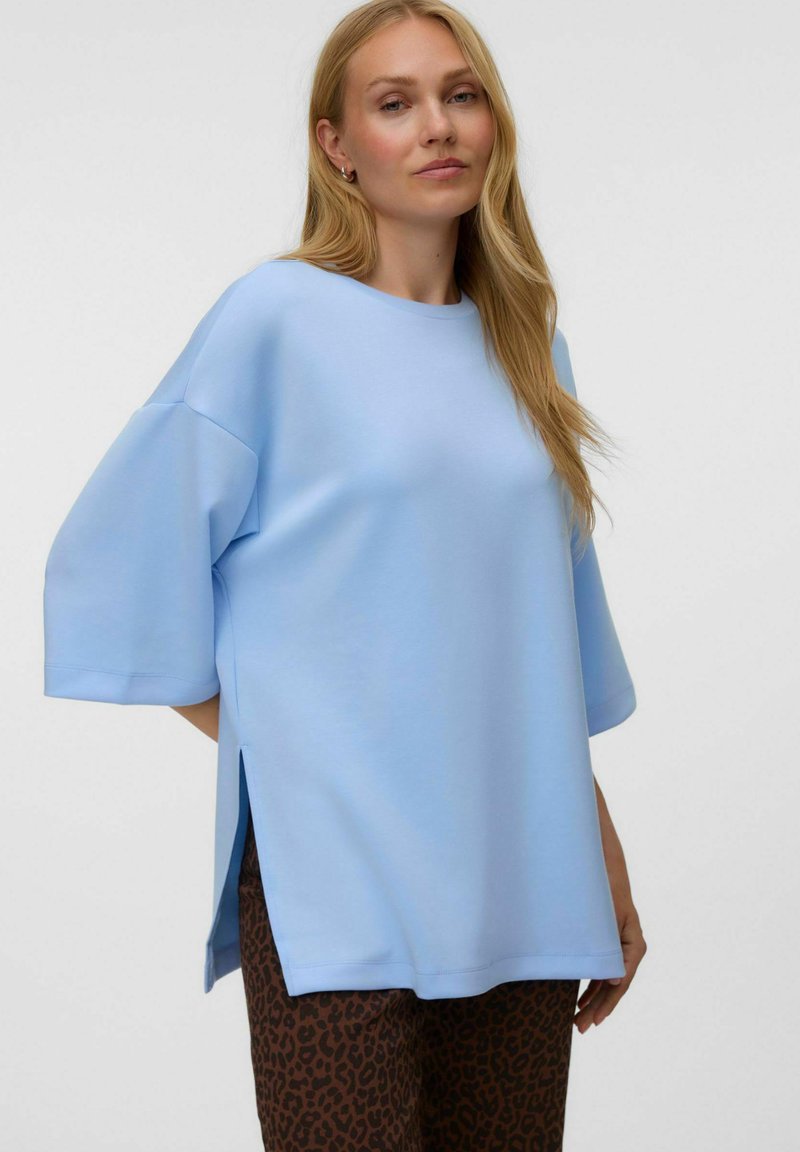 Femme aux longs cheveux blonds porte un haut ample bleu clair avec des manches jusqu'aux coudes et un pantalon marron imprimé léopard sur un fond uni.