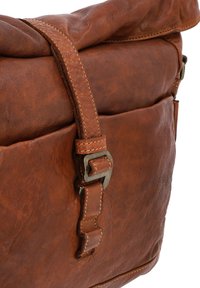 Brūna ādas kurjera soma ar teksturētu virsmu, ruļļu apakšējo daļu, regulējamu siksnu un metāla sprādzi sānos.