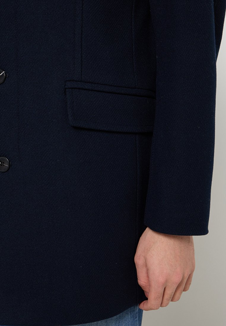 Marineblauwe wollen jas met een gestructureerd ontwerp, voorzien van een enkele zak, knoopsluiting en een textuurafwerking, gedragen over een lichtblauw shirt.