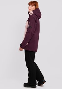 Purple and light pink waterproof jacket with a hood, zippered pockets, and adjustable cuffs, worn over black pants and footwear.
