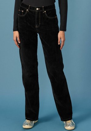 Czarne spodnie kordrowe o prostym kroju nogawek, z kieszeniami i kontrastującym przeszyciem. Zestawione z szarymi sneakerami, na niebieskim tle.