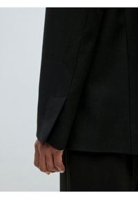 Zwarte op maat gemaakte blazer met een gestructureerd ontwerp, scherpe revers en een enkele vent. Gladde textuur en aansluitende mouwen met een nette stikseldetail.