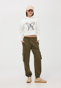 Felpa bianca cropped con grafica di una farfalla, abbinata a pantaloni cargo verde oliva e sneakers beige; presenta polsini elasticizzati e tasche laterali.