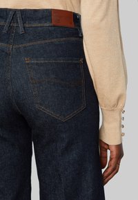 Närbild av en person som bär mörkblå jeans med orange sömnad och en beige långärmad tröja med pärlknappar på ärmen.