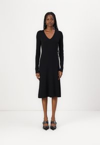 Black ribbed dress with a V-neck and long sleeves, reaching mid-calf, paired with black studded heeled shoes. Smooth, fitted texture.
