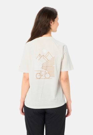 Vrouw van achteren gezien die een beige T-shirt draagt met een minimalistische afbeelding van een berg, zon, weg en fiets op de rug, gecombineerd met een zwarte broek.