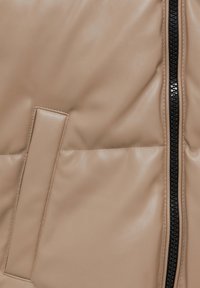 Section de veste matelassée beige avec une fermeture éclair noire verticale et une poche horizontale sur le côté gauche.