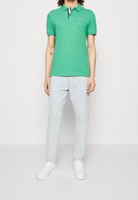 Camisa polo verde con cuello y mangas cortas, con un pequeño logo. Combinada con jeans azul claro y zapatillas blancas.