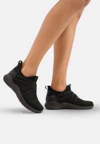 Schwarze Sportschuhe mit Obermaterial aus Mesh, strukturierter Sockenpassform, elastischen Riemen und erhöhter profilierter Sohle. Ideal für Komfort und Atmungsaktivität.