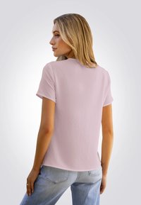 Mujer rubia con camiseta de manga corta rosa claro y jeans azul claro posa de espaldas, mirando por encima del hombro contra un fondo liso.