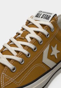 Καφέ αθλητικό παπούτσι Converse All Star με λευκά κορδόνια, λευκό αστέρι και λογότυπο σχήματος βέλους, και αντιθετικές λεπτομέρειες στη σόλα σε λευκό και μαύρο χρώμα.