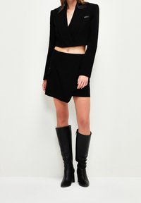 Chaqueta negra corta con escote en V profundo y detalles de cuero, combinada con una falda corta negra. Botas altas negras completan el conjunto.