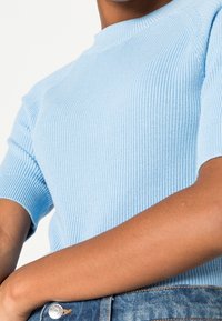 Jasnoniebieski prążkowany krótki top z dzianiny z wysokim kołnierzem, wykonany z teksturowanej tkaniny o luźnym kroju, noszony z dżinsowymi spodniami.