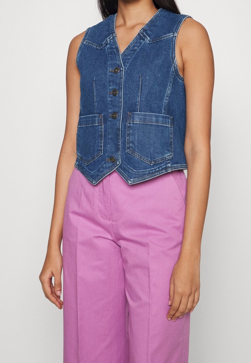 Gilet en jean bleu sans manches avec boutons et poches avant, porté avec un pantalon rose taille haute à jambes larges sur une personne debout.
