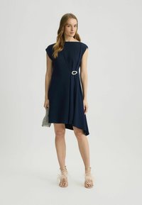 Tengerész kék, aszimmetrikus ruha, kapucnis ujjakkal és díszes, fehér, kör alakú övvel. A modellen egy texturált ezüst színű Clutch, valamint tollas szandál található.