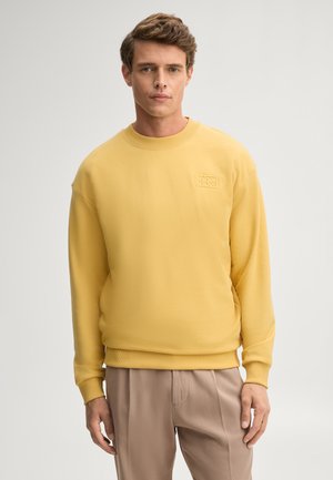 Ein junger Mann mit kurzen braunen Haaren trägt ein gelbes Sweatshirt und beige Hose, steht vor einem schlichten hellen Hintergrund.
