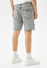 Mies, joka käyttää vaaleanharmaita kuluneita farkkushortseja, valkoisia sukkia ja valkoisia tennareita, seisoo valkoista taustaa vasten.