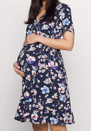 Gravid kvinde iført en marineblå omslagskjole med lyserødt og blåt blomsterprint, som blidt holder sin babybule med begge hænder.